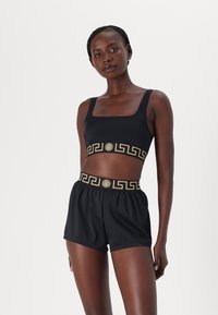 Must mustatud sportlik crop-top laia rihmaga ja kuldse mustriga riba. Kombineeritud mustade lühikeste pükstega, millel on kuldse aktsendiga elastne vöökoht.