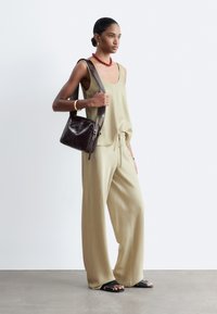 Femme portant un top beige sans manches et un pantalon à jambes larges, portant un sac à bandoulière en cuir brun foncé, accessoirisée d'un collier rouge et d'un bracelet en or.