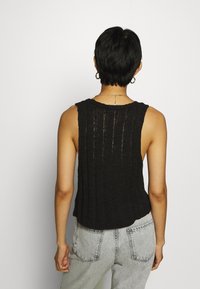 Black knit sleeveless top with vertical texture, loose design, and round neckline. Paired with light gray jeans, viewed from the back.
