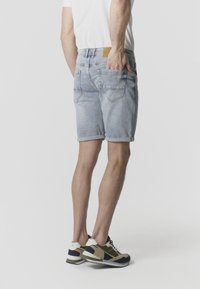 Személy áll, kezét a zsebében tartva, világos, visszahajtott farmer rövidnadrágot, fehér pólót és bézs-fekete tornacipőt visel, egyszerű háttér előtt.