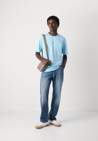 Νεαρός άνδρας φορώντας ανοιχτό μπλε T-shirt, μπλε τζιν, λευκά αθλητικά παπούτσια και τσάντα χιαστί, στέκεται μπροστά σε λευκό φόντο.