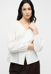 Λευκή μπλούζα από ελαφρύ ύφασμα με λαιμόκοψη V, φουσκωτά μανίκια και χαλαρή εφαρμογή με ευθεία τελείωμα. Χωρίς ορατά σχέδια ή διακοσμήσεις.