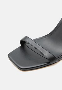 Μαύρο δερμάτινο σανδάλι με τετράγωνη μύτη και μία λεπτή λωρίδα μπροστά, απεικονίζεται σε κοντινή λήψη με λευκό φόντο.