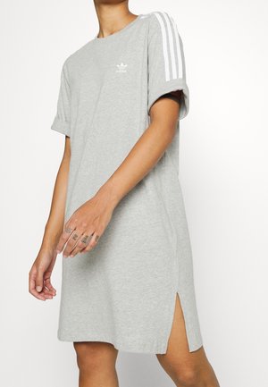 Personne portant une robe t-shirt Adidas grise avec des bandes blanches sur les manches et une fente latérale, montrant des doigts tatoués.
