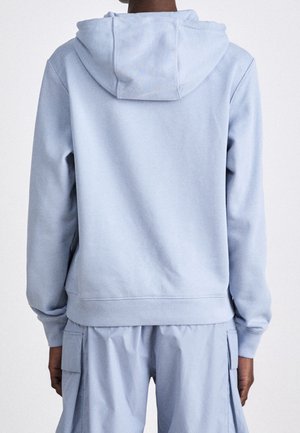 Personne portant un sweat à capuche bleu clair et un pantalon cargo assorti, vue de dos sur un fond uni.