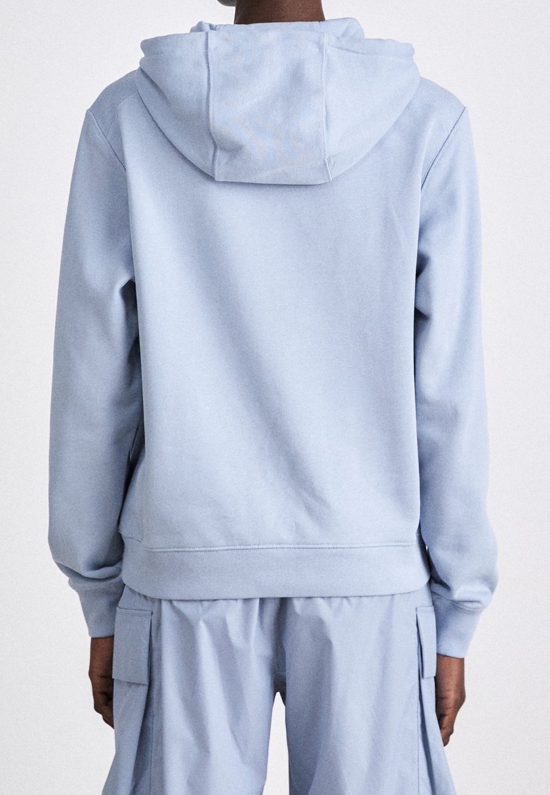 Personne portant un sweat à capuche bleu clair et un pantalon cargo assorti, vue de dos sur un fond uni.