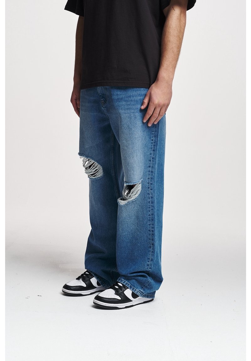 Man wearing loose blue ripped jeans and black-and-white sneakers, standing against a plain light background.