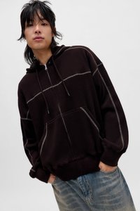 Bruine hoodie met ritssluiting, textuurstof en contrasterende lichte stiksels, voorzien van een capuchon en zakken aan de voorkant, gedragen over een blauwe spijkerbroek.