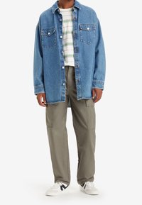 Lichtblauw denim overhemd met twee borstzakken en knoopsluiting. Gecombineerd met olijfgroene cargobroek en witte sneakers met zwarte accenten.