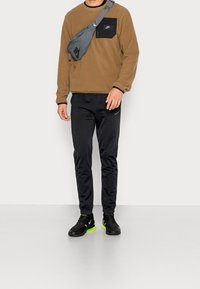 Brun fleece-tröja med svart bröstficka, svarta sportbyxor och svarta skor med neongröna detaljer. Axelväska bärs tvärs över kroppen.