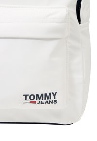 Biała torba ma gładką powierzchnię z dużą przednią kieszenią. Logo "TOMMY JEANS" w czerni z czerwonymi i niebieskimi akcentami na dole.