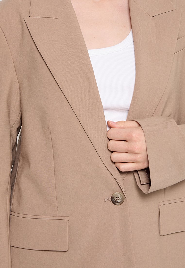Blazer beige chiaro realizzato in tessuto liscio, con un unico bottone in vita, rever affilati e due tasche frontali. Maniche lunghe e larghe.