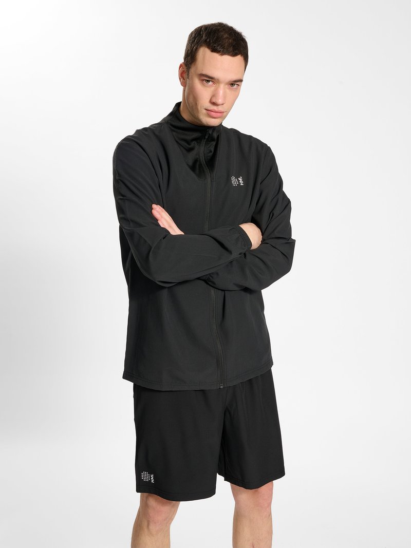 Homme portant une veste noire zippée et un short noir, debout les bras croisés devant un fond clair et uni.