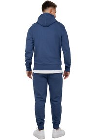 Marineblauwe hoodie met een buidelzak, verstelbare trekkoordcapuchon en bijpassende joggers met elastische boorden, gedragen met witte sneakers.