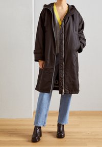 Manteau long noir avec fermeture éclair à l'avant, capuche, et poches latérales, porté par-dessus un haut gris et un jean large bleu clair, associé à des bottes noires.