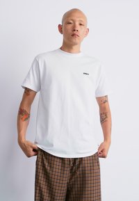 White cotton t-shirt with crew neck, short sleeves, and a small black "OBEY" logo on the chest, paired with brown checked shorts.