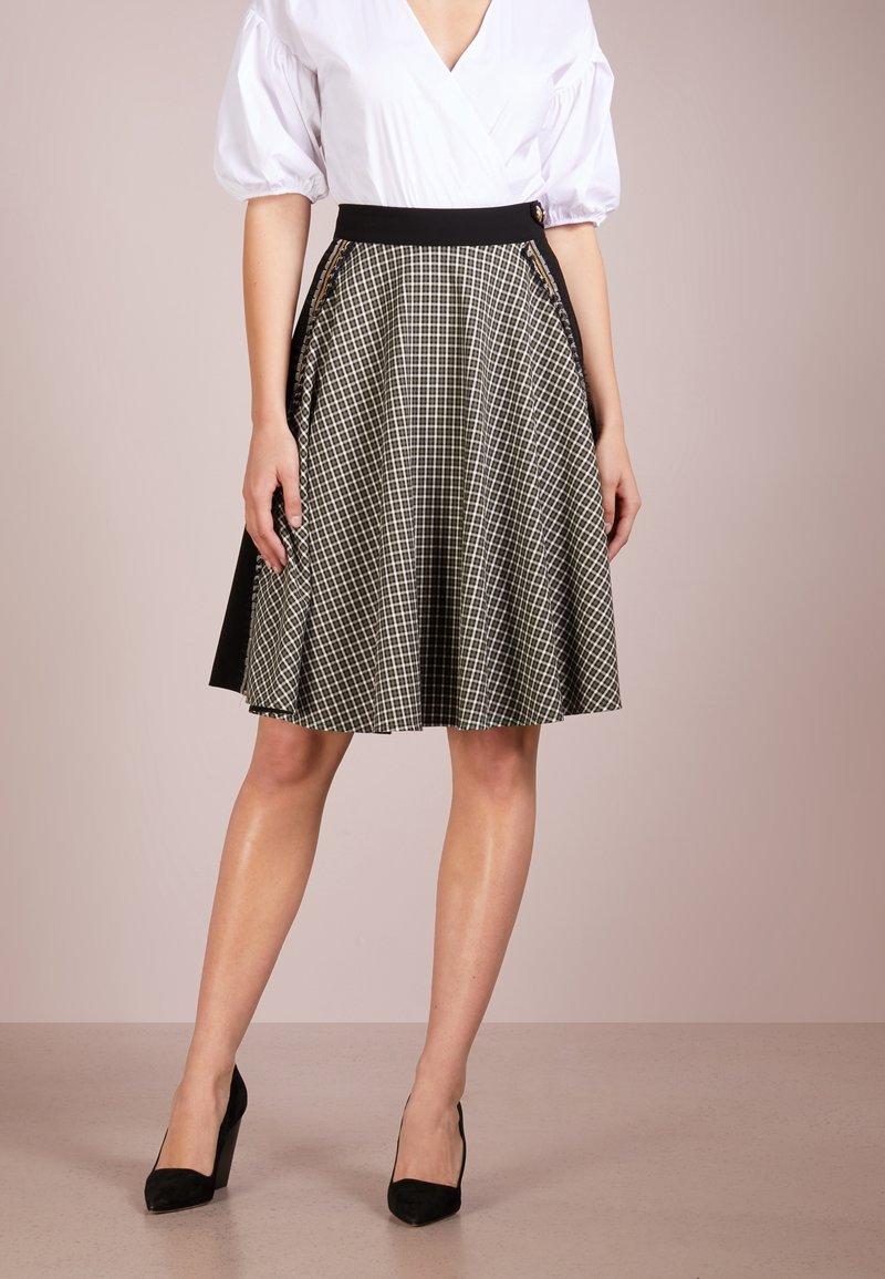 Jupe à carreaux noir et blanc avec une taille ajustée, fermeture éclair latérale et design plissé. Portée avec une blouse blanche et des talons noirs.