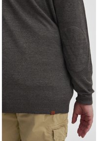 Suéter gris oscuro con dobladillo y puños acanalados, con parches en los codos, combinado con pantalones cargo de color claro. Etiqueta de cuero visible.