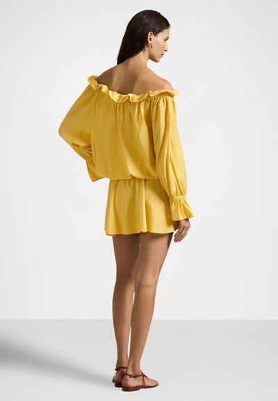 Γυναίκα σε προφίλ φορώντας κίτρινο φόρεμα με γυμνούς ώμους, με βολάν στο ντεκολτέ και μακριά μανίκια, συνδυασμένο με καφέ φλατ σανδάλια.