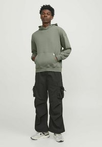Felpa verde oliva con tasca frontale e cappuccio regolabile, abbinata a pantaloni cargo neri con multiple tasche laterali. Indossati con sneakers grigie.