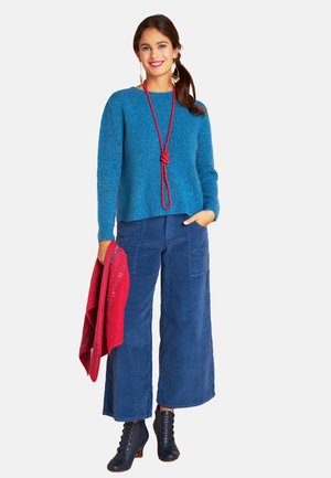 Blauer Strickpullover, weite marineblaue Cordhosen und schwarze Stiefel mit Absatz. Ein roter Schal und eine lange Halskette mit roten Perlen setzen farbige Akzente.