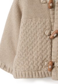 Cardigan beige lavorato a maglia con motivi testurizzati, bordo a costine e bottoni in legno fissati con anelli di filato.