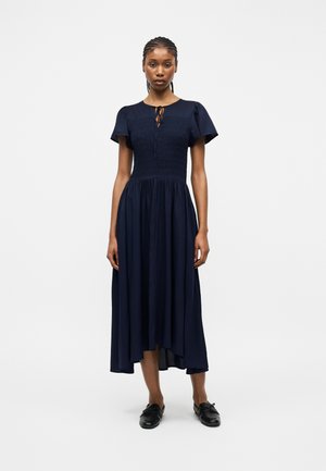 Frau steht und trägt ein marineblaues Midikleid mit kurzen Ärmeln, gerafftem Oberteil, gebundenem Halsausschnitt und schwarzen Loafers vor weißem Hintergrund.