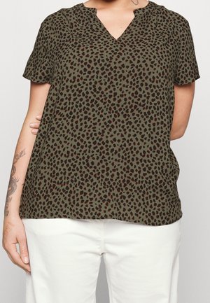 Top court à manches courtes vert olive avec imprimé léopard noir et marron, porté avec un pantalon blanc, montrant un avant-bras tatoué.