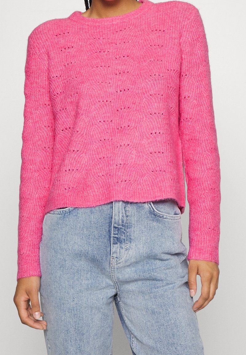 Różowy sweter na drutach z wzorem fal, okrągłym dekoltem i ściągaczami na rękawach, zestawiony z jasnoniebieskimi jeansami.