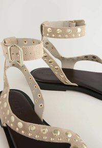 Beige suède sandalen met gouden ronde studs. Voorzien van een verstelbare enkelriem en een vlakke zwarte zool. Open teen ontwerp met een minimalistische stijl.
