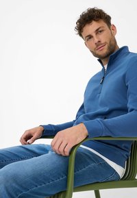 Felpa con zip blu, maniche lunghe, dotata di un colletto alto e rivestimento interno bianco a contrasto, abbinata a jeans azzurri.
