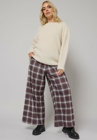 Pull en tricot crème à manches longues, associé à un pantalon à carreaux bordeaux à jambes larges et des bottes à talons épais noires. Texture lisse et motifs distincts.