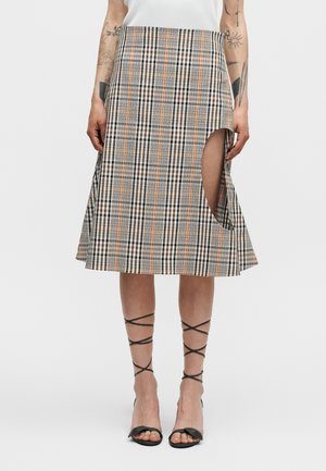 Kobieta ubrana w brązową i pomarańczową midi spódnicę w kratę z wysokim rozcięciem po boku, zestawioną z czarnymi sandałami na wysokim obcasie z paskami i białym topem.