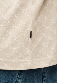 Μπεζ υφασμάτινη υφή με διακριτικό μοτίβο διαμαντιού και μικρή ετικέτα λογότυπου πάνω από μπλε τζιν.