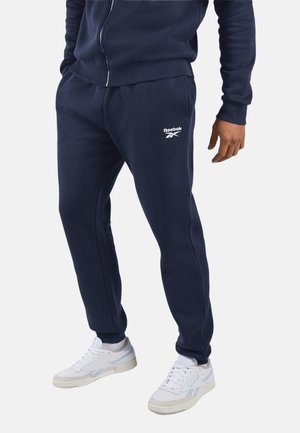 Personne portant un jogging Reebok bleu marine et une veste zippée assortie, baskets blanches Reebok avec des touches bleu clair visibles.