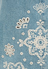 Υφασμα από ανοιχτό γαλάζιο τζιν με λευκά κεντήματα λουλουδιών και γεωμετρικών σχεδίων σε διάφορα σχήματα και μεγέθη.