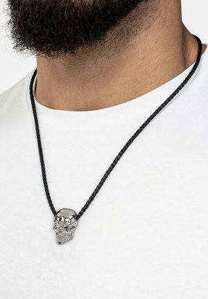 Silberner Totenkopf-Anhänger an einer schwarzen geflochtenen Schnur-Halskette, getragen über einem weißen T-Shirt. Verfügt über filigrane Details auf dem Totenkopf.