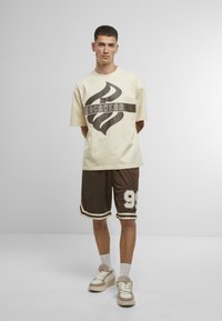 Chemise beige à manches courtes avec un logo abstrait noir, associée à un short marron arborant un "98" blanc et des rayures décoratives. Baskets beiges.