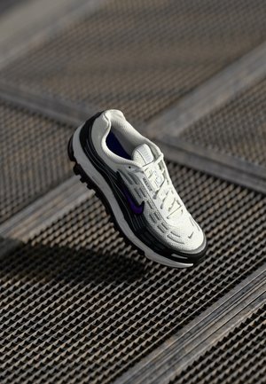 Biało-czarny but do biegania Nike z fioletowym logo unoszący się nad teksturowaną metalową powierzchnią.
