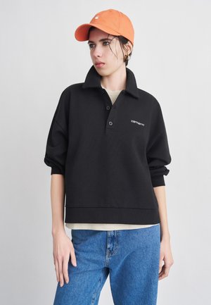 Giovane con indosso un berretto arancione, una felpa nera con colletto, una camicia beige sotto e jeans blu, su uno sfondo chiaro e semplice.