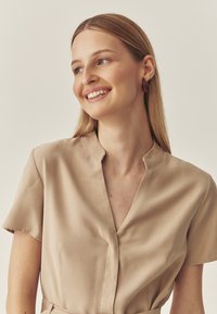 Beige Bluse mit kurzen Ärmeln und V-Ausschnitt, aus glattem Stoff und leicht taillierter Passform. Das Modell trägt bordeauxfarbene Ohrhänger.