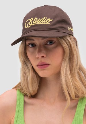 Joven mujer con cabello largo y rubio que lleva una gorra marrón con texto amarillo "studio" y una camiseta sin mangas de color verde claro, sobre un fondo liso.