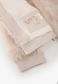 Giacca imbottita in beige chiaro con dettagli in pelliccia sintetica, dotata di un patch con logo dorato e chiusura lampo dorata. Texture liscia con morbidi accenti in pelliccia.