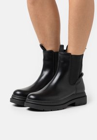 Svarta läderankelbootar med elastiska sidopaneler, rundad tå, dragflikar och en tjock gummisula. Slät yta och enkel design.