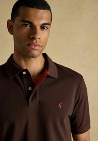 Gaiši brūns polo krekls ar sarkanu apkakles akcentu un baltām pogām. Uz kreisās puses atrodas mazs logo. Gluda tekstūra, klasiskā piegriezuma.