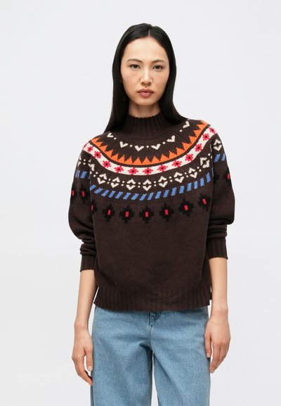 Femme portant un pull en maille marron foncé avec des motifs géométriques colorés et un jean bleu clair, debout devant un fond blanc uni.