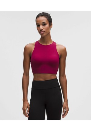 Donna che indossa un top corto senza maniche di colore rosa scuro e leggings neri a vita alta, in piedi davanti a uno sfondo chiaro e semplice.