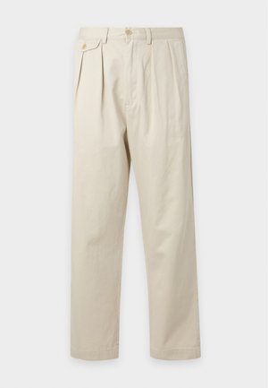 Jeune homme portant une casquette bleu marine, un gilet matelassé vert, une chemise à carreaux violets, un pantalon beige et des mocassins beiges, debout devant un fond blanc uni.