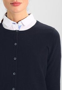 Detailní záběr na výstřih ženy, která má na sobě tmavě modrý kardigan přes bílou košilovou blůzu, s viditelnými knoflíky a jemným výrazem.