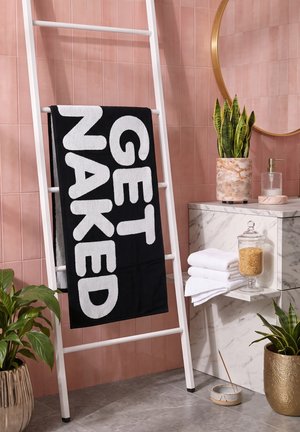 Serviette noire avec le texte "GET NAKED" suspendue à une échelle blanche contre un mur carrelé rose, à côté de plantes et d'un lavabo en marbre avec des articles de toilette.